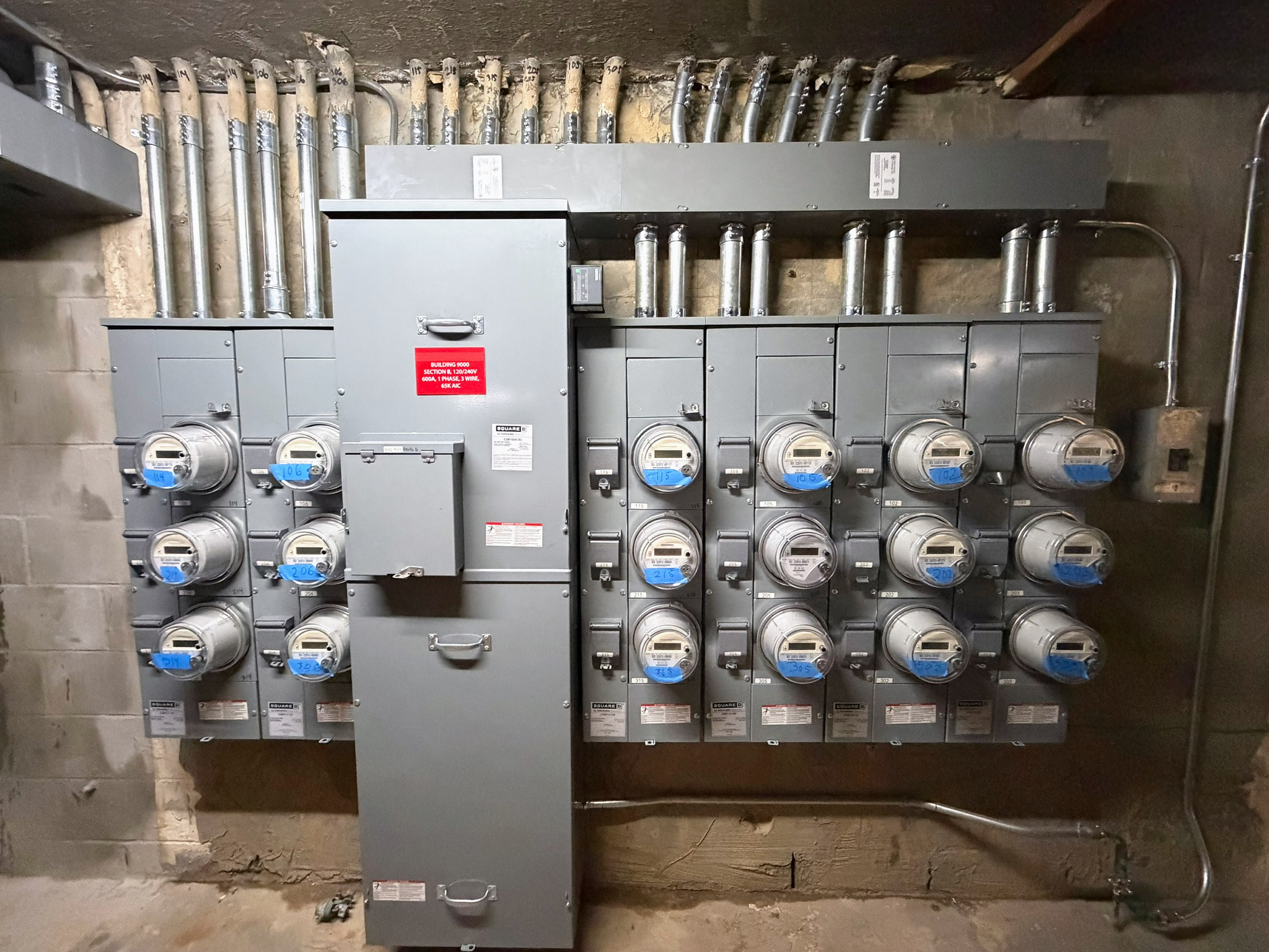 Summertime Section B electrical meter bank with a central service cabinet, labeled meters and overhead metal conduit.