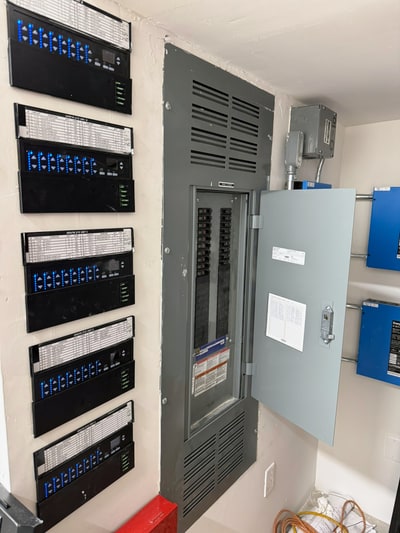 Open circuit breaker panel beside stacked lighting controls and blue control enclosures in an electrical room.