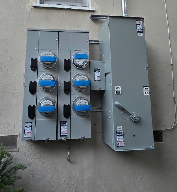 Six electric meters arranged in two columns beside a large exterior service disconnect, with metal conduit rising along a stucco wall.