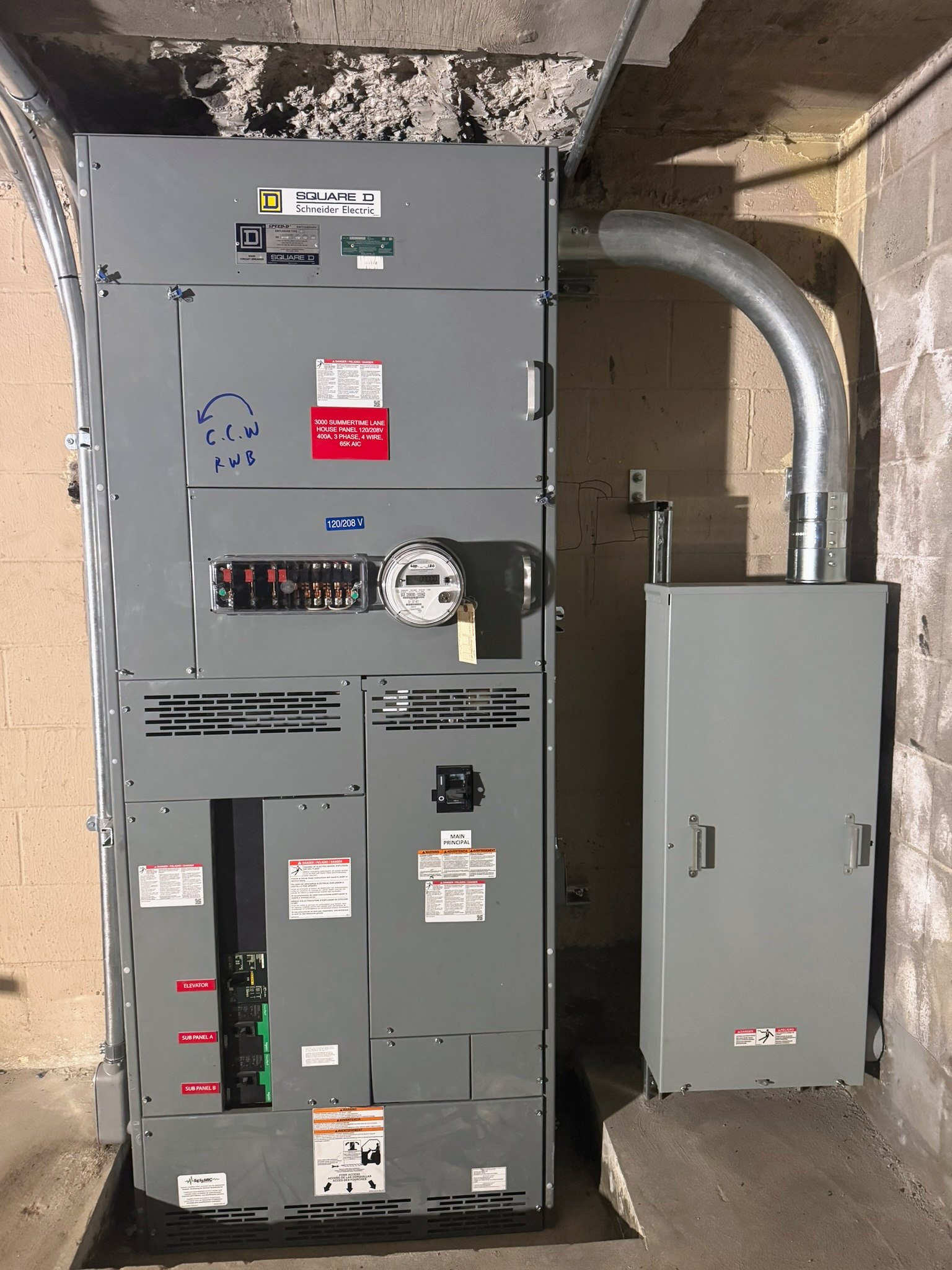 Close view of the Summertime house service panel with electric meter, labeled breakers and a large curved conduit to an adjacent enclosure.
